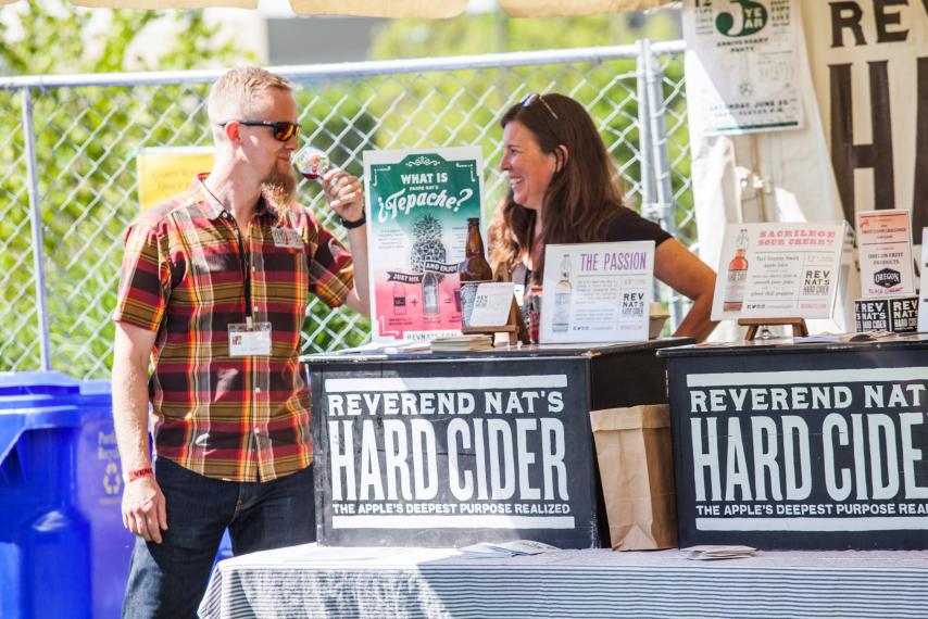 A man and woman standing next to a table selling hard cider.
